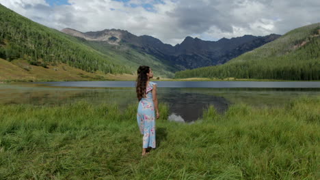 Aerial-cinematic-drone-following-female-women-model-actress-cute-dress-walking-toward-arms-wide-Piney-Lake-Ranch-Vail-Beaver-Creek-Colorado-Gore-Range-mountain-landscape-summer-afternoon-rain-clouds