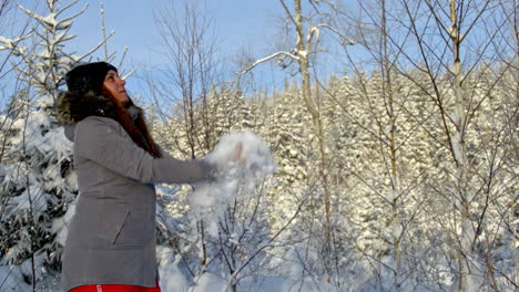 Woman-tosses-handful-of-snow-up-into-air-in-front-of-forest,-snow-flurries-fly