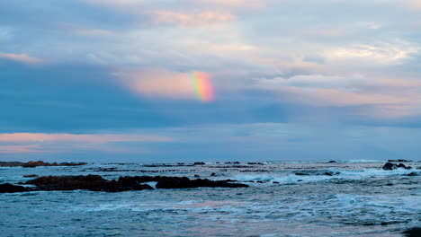 Blick-über-Wellen-Und-Küstenfelsen-Mit-Teilweise-Regenbogen-In-Den-Sonnenuntergangswolken