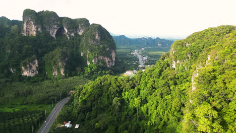 Ascending-drone-shot-of-traffic-on-road-between-green-mountains-of-Krabi-Province-at-sunset,-Thailand