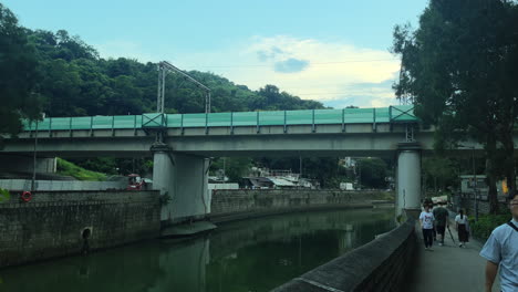 Static-shot-of-people-walking-on-a-busy-walkway-next-to-a-slow-river-going-under-a-railway-bridge