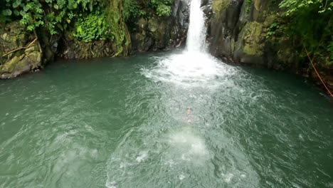 Young-Adult-man-at-Kroya-Waterfall-cliff-jump-fearless-off-a-diving-Platform-headfirst-into-natural-Pool-amid-lush-rock-walls-and-forest-of-Aling-Aling,-Bali