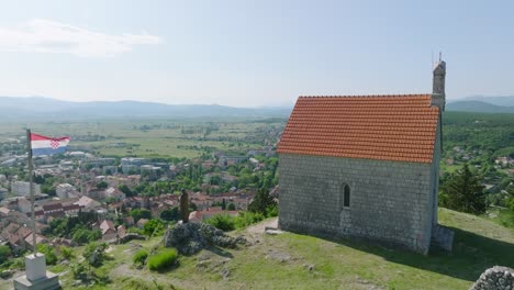 Croatian-flag-by-Our-Lady-of-Sinj-Church-on-hill-by-town,-aerial-pan