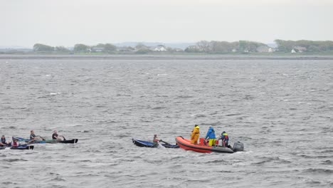 Medium-view-of-mixed-currach-racers-rowing-through-ocean-with-orange-inflatable-boat-following