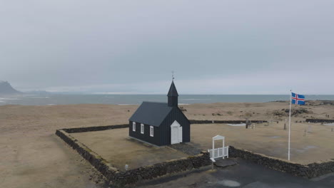 Drone-Shot-of-Church-in-Búðir,-Iceland,-Black-Wooden-Building-and-National-Flag-by-the-Sea
