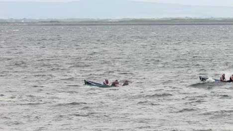 Tracking-pan-follows-currach-boats-racing-through-open-waters,-orange-safety-boat-following-behind