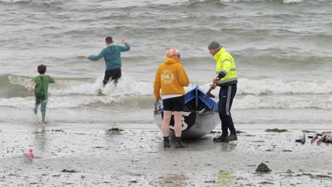 Safety-officer-carries-oar-to-currach-boat-on-shore-as-young-boys-play-in-chilly-cold-water