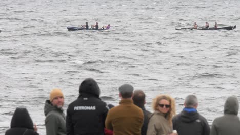 Spectators-watch-in-foreground-as-currach-boats-row-in-front-in-slow-motion