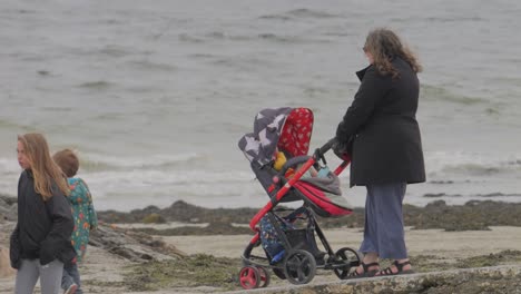 Woman-pushes-stroller-as-young-boy-child-leads-the-way-down-to-the-beach,-ireland