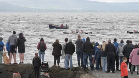 People-stand-on-sea-wall-jetty-watching-currach-boats-race-in-galway-bay-ireland