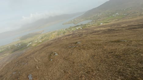 Drohne-Fliegt-über-Einen-Bergrücken-Durch-Tief-Hängende-Wolken-In-Richtung-Eines-Kleinen-Dorfes-Am-Atlantischen-Ozean,-Irland