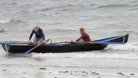 Couple-pushes-to-launch-currach-row-boat-into-ocean-through-breaking-waves-on-shore