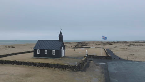 Vista-Aérea-De-La-Iglesia-Negra-De-Búðakirkja,-Punto-De-Referencia-De-Islandia,-Exterior-Y-Bandera-Nacional