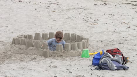 Young-boy-digs-deep-hole-and-creates-sand-castle-using-buckets-spread-around-him-at-beach