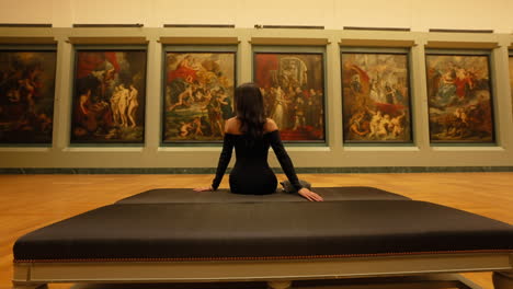 Beautiful-elegant-woman-sitting-down-in-front-of-historical-paintings,-in-an-art-gallery-at-the-Louvre-Museum,-Paris,-France