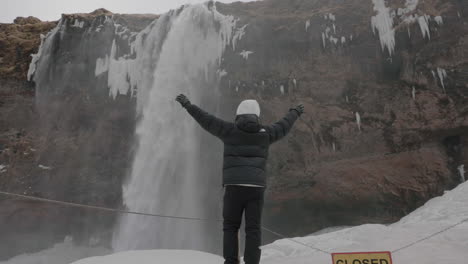 Rücken-Eines-Mannes-In-Warmer-Winterkleidung,-Der-Auf-Schnee-Unter-Einem-Wasserfall-Auf-Island-Läuft