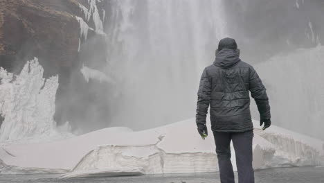 Back-View-of-Man-Walking-on-Ice-Under-Waterfall-on-Cold-Winter-Day,-Slow-Motion