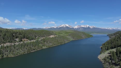 Aerial-view-over-the-lake-surrounded-by-forest,-snowy-mountain-range-of-Colorado-Mountains
