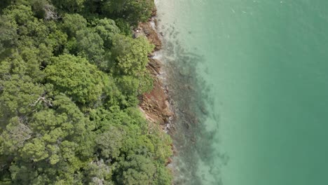 Warm-water-laps-at-rock-shoreline-and-tropical-sand-beach,-aerial-view