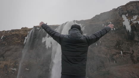 Back-of-Man-Walking-in-Snow-in-Warm-Winter-Clothes-and-Raising-Arms-in-Front-of-Waterfall,-Slow-Motion