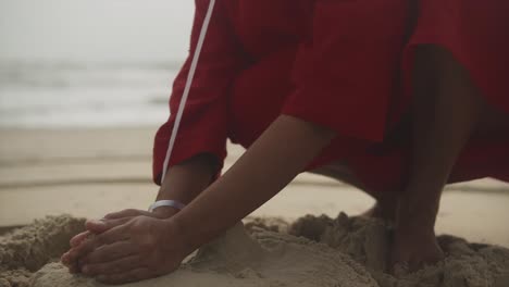 Close-up-shot-of-hands-of-a-girl-forming-a-sand-castle-on-the-beach-with-his-hand-in-red-dress---close-up,-slow-motion-shot