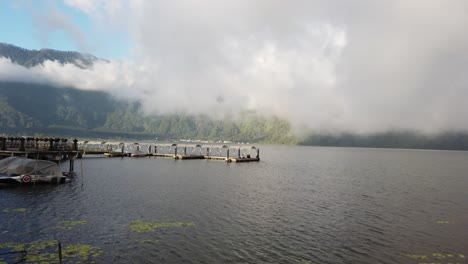Panoramic-view-of-Beratan-Lake-in-Bali,-Indonesia-with-a-light-breeze,-fog-clearing-up-and-the-sun-trying-to-break-through-revealing-mountains-in-the-background