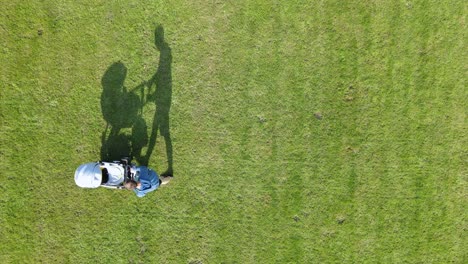 Dad-father-pushing-a-baby-stroller-with-his-son-during-a-sunny-day-of-summer-in-green-field-park,-aerial-top-down