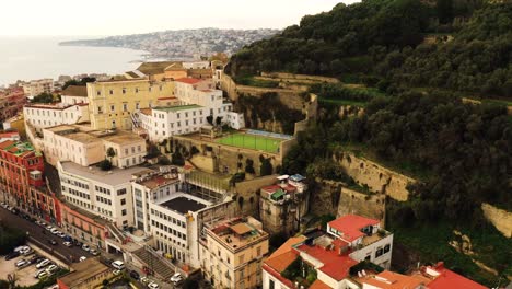 Aerial-orbiting-shot-showing-old-buildings-with-soccer-field-located-on-hill-in-Naples-City-during-golden-hour---Coastline-and-sea-in-background