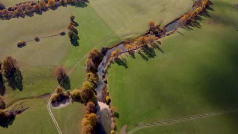 Natural-river-flows-through-Slovenian-grassland,-aerial-tilt-up-autumn-forest