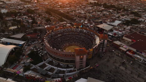Aerial-view-away-from-the-Plaza-de-Toros-stadium,-sunset-in-Aguascalientes,-Mexico---reverse,-tilt,-drone-shot