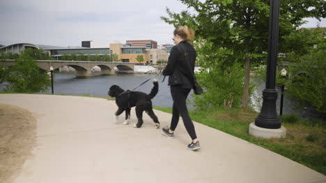 Handheld-shot-of-a-young-woman-walking-her-dog-along-a-river-front-path