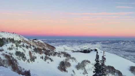 Tilt-up-shot-of-a-stunning-winter-landscape-scene-looking-down-at-a-cloudy-snow-covered-valley-during-a-vibrant-colorful-sunset-from-the-summit-of-a-ski-resort-in-the-Rocky-Mountains-of-Utah