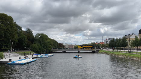 Malmö-Kanal:-Menschen,-Die-Mit-Einem-Fahrradboot,-Einem-Bus-Und-Einem-Lastwagen-über-Eine-Brücke-Fahren,-Bäume-An-Der-Seite-Und-Gebäude-Im-Hintergrund
