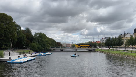 Malmö-canal:-People-riding-a-cycle-boat,-a-bus-and-a-truck-crossing-a-bridge,-trees-on-the-side-and-buildings-in-the-background