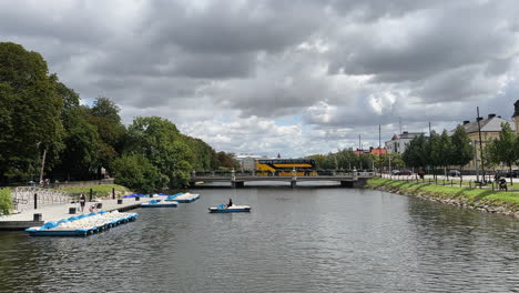 Overview-of-Malmö-canal:-People-riding-a-cycle-boat,-a-bus-and-a-truck-crossing-a-bridge,-trees-on-the-side-and-buildings-in-the-background