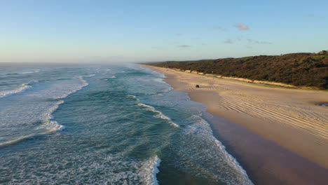 Aerial-view-following-a-car-driving-on-the-K'gari-beach,-golden-hour-in-Australia