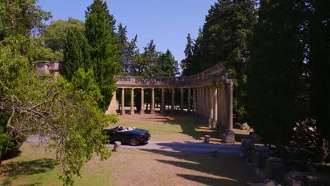 In-the-garden,-there-is-a-stone-structure-with-columns,-and-a-black-car-is-driving-along-the-road