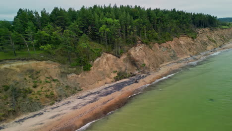 Aerial-of-green-Baltic-sea-at-Jurkalne-bluffs,-waves-erode-the-forest-near-the-beach