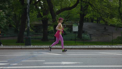 Slow-motion-of-blonde-woman-with-red-hat-holding-phone-jogging-outdoors-in-New-York-City-park