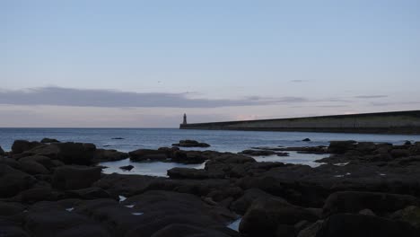 Wide-angle-fixed-establishing-clip-of-long-sea-pier-with-lighthouse-at-the-end,-and-dark-rocks-and-rockpools-in-the-foreground