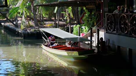 Sentado-Junto-A-Las-Escaleras,-El-Barquero-Espera-A-Sus-Pasajeros-Que-Están-Comprando-Recuerdos-En-Una-De-Las-Tiendas-Más-Populares-Del-Mercado-Flotante-Damnoen-Saduak-En-Tailandia