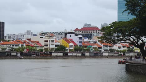 Un-Barco-Turístico-Se-Mueve-Lentamente-A-Lo-Largo-Del-Río-Singapur-Con-Las-Coloridas-Casas-Que-Bordean-El-Muelle-De-Barcos-Al-Fondo,-Singapur