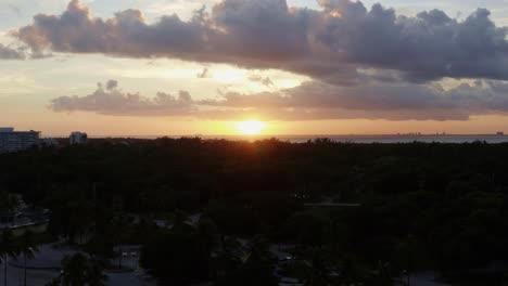 Dolly-in-rising-aerial-drone-shot-of-a-stunning-yellow-golden-ocean-sunset-with-tropical-greenery-below-from-Crandon-Park-in-Key-Biscayne-outside-of-Miami,-Florida-on-a-warm-sunny-summer-evening