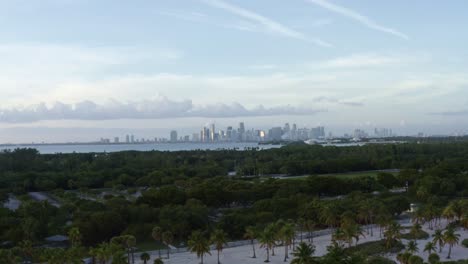 Dolly-out-aerial-drone-shot-of-the-tropical-beach-surrounded-by-palm-trees-on-Crandon-Park-in-Key-Biscayne-with-the-skyline-of-Miami,-Florida-in-the-distance-on-a-sunny-summer-evening