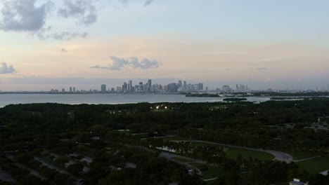 Dolly-out-descending-aerial-drone-shot-of-the-tropical-beach-surrounded-by-palm-trees-on-Crandon-Park-in-Key-Biscayne-with-the-skyline-of-Miami,-Florida-in-the-distance-on-a-sunny-summer-evening