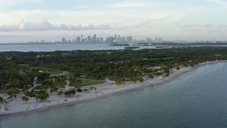 Tilting-up-left-trucking-aerial-drone-shot-of-the-tropical-beach-surrounded-by-palm-trees-on-Crandon-Park-in-Key-Biscayne-with-the-skyline-of-Miami,-Florida-in-the-distance-on-a-sunny-summer-evening