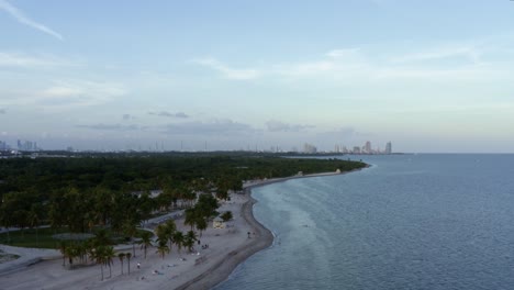 Dolly-out-aerial-drone-shot-of-the-beautiful-tropical-beach-surrounded-by-palm-trees-on-Crandon-Park-in-Key-Biscayne-outside-of-Miami,-Florida-on-a-warm-sunny-summer-evening
