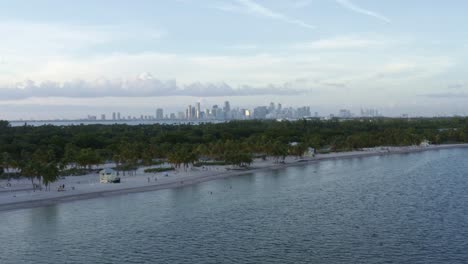 Dolly-in-tilting-up-aerial-drone-shot-of-the-tropical-beach-surrounded-by-palm-trees-on-Crandon-Park-in-Key-Biscayne-with-the-skyline-of-Miami,-Florida-in-the-distance-on-a-sunny-summer-evening