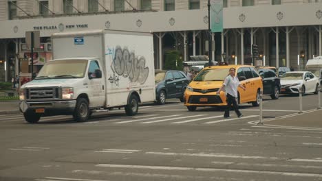 Yellow-taxi-cabs-and-transport-van-with-graffiti-drive-through-intersection-fronting-plaza-hotel
