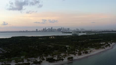 Dolly-out-descending-aerial-drone-shot-of-the-tropical-beach-surrounded-by-palm-trees-on-Crandon-Park-in-Key-Biscayne-with-the-skyline-of-Miami,-Florida-in-the-distance-on-a-sunny-summer-evening